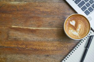 Cozy workspace featuring a cappuccino with latte art, open notebook, and laptop on a wooden desk.